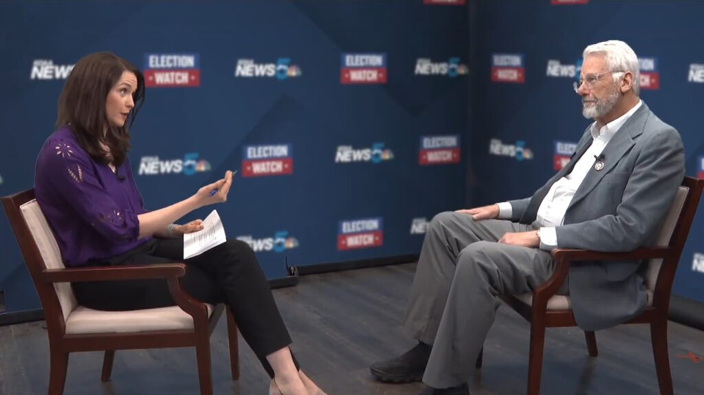 Interviewer and man sitting apart with "Election Watch" backdrop